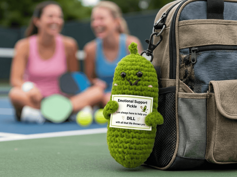 Emotional Support Pickle crocheted green pickle with a care tag sitting on a pickleball bag beside two players