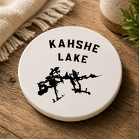 Round ceramic Kahshe Lake coaster with black lake silhouette on a wooden table beside a fringed blanket