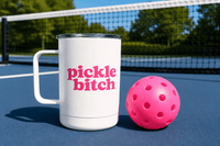 White 12 oz insulated pickleball travel mug that says pickle bitch beside a pink pickleball on a court