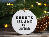 Round ceramic Courts Island PEI coordinates ornament hanging beside evergreen branches, pinecones and Christmas lights